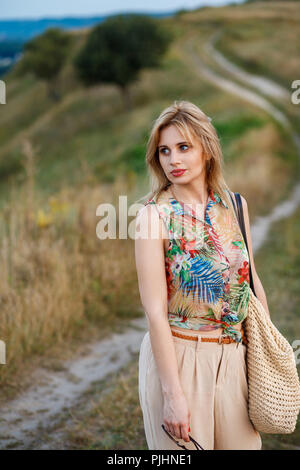 Young beautiful thoughtful woman in tropical blouse and beige galligaskins with wicked bag standing on village road and looking aside. Outdoor shot, n Stock Photo