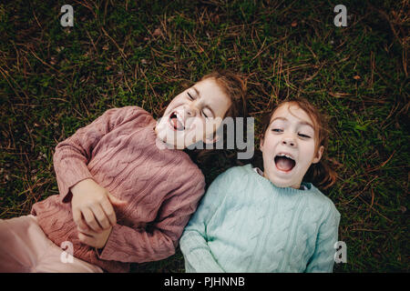 Top view of two little girls lying on green grass and laughing. Twins sisters enjoying outdoors at the park. Stock Photo