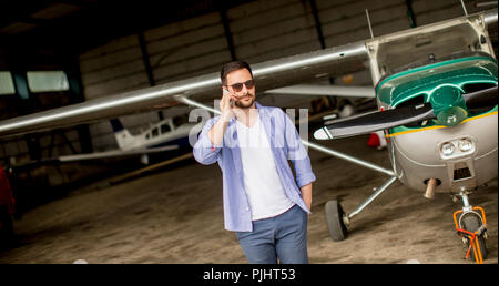 Young handsome young pilot checking airplane in the hangar and using mobile phone Stock Photo