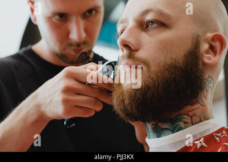 A young handsome bearded man in a barbershop. The barber brushes his hair and cuts it with an electric trimmer. Stock Photo