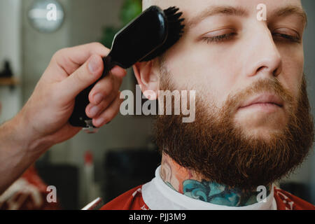 A handsome bearded skinhead man in a barbershop. The barber swipes off the cut hair with a brush. Stock Photo