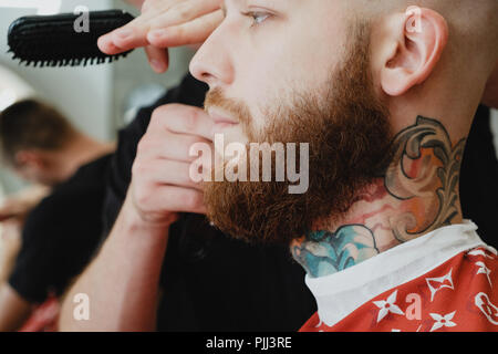 A young handsome bearded man in a barbershop. The barber brushes his hair and cuts it with an electric trimmer. Stock Photo
