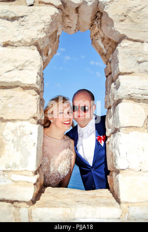 Wedding. Happy couple day wedding. Beautiful bride and groom at the beach. Cheerful married couple standing and posing on the stone wall window. Stock Photo