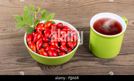 Rosehip tea. Red briar fruit pile in bowl. Wood background. Rosa canina. Smoking medicinal drink in mug. Fresh ripe wild hips. Twig with green leaves. Stock Photo