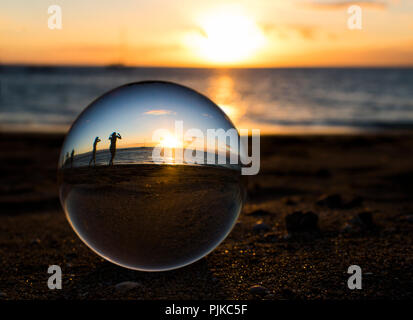 Glass Ball Captures Sunset on Beach with People in Silhouette Stock Photo