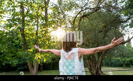 Young woman celebrating the morning sun and spring season as she stands outdoors in the garden at sunrise with outspread arms, rear view. Stock Photo