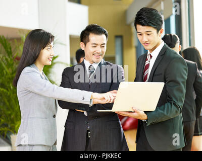 two asian businessmen and a businesswoman discussing business using laptop computer in company lobby. Stock Photo