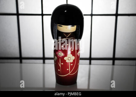 Japanese wooden doll. Small Geisha toy wearing a kimono with cherry blossom patters on a white surface and paper divider in the background. Stock Photo