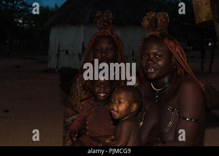 Two Himba women sit in the dark with their children on their laps at the fire, smiling at the camera Stock Photo