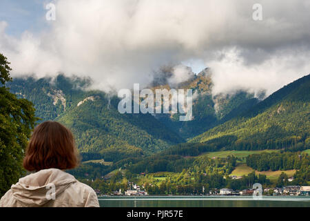 View of the lake near St. Wolfgang with a woman looking at the Alps mountains in the background, under a blue sky with clouds on a sunny day Stock Photo