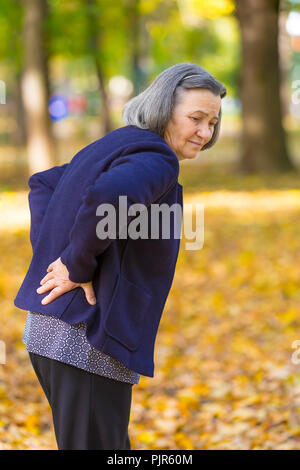 Elderly woman with lower back pain in autumn city park. Unhappy senior woman while feels pain from backache walking in autumn park. Stock Photo