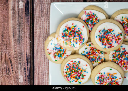 Vanilla frosted sugar cookies with rainbow colored sprinkles. Plated view, wooden background Stock Photo