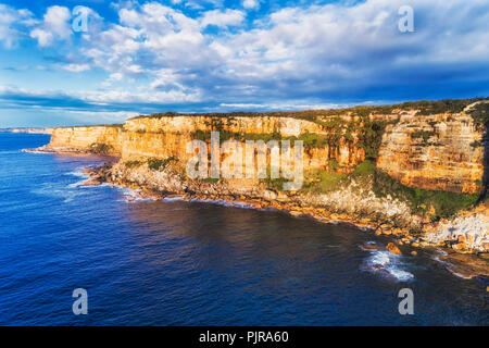 Tall eroded rugged sandstone cliffs of Sydney's North head at the entrance to Sydney harbour hit by endless waves of Pacific ocean lit by warm morning