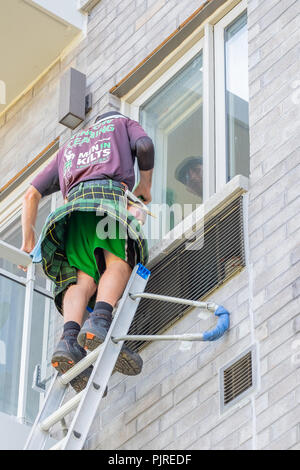 Man on a ladder wearing a kilt flapping in the wind washes windows in an apartment complex. Stock Photo