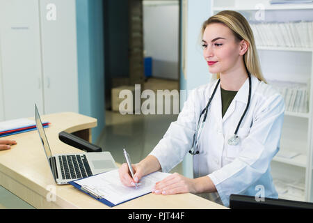 Smiling young female doctor working at the clinic reception, she is using a computer and writing medical reports Stock Photo