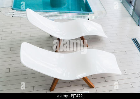 Empty two white lounge chairs inside of tiled room near swimming pool. Nobody in spa room. Deck chair for clients relaxation after spa procedures, bath house and swimming in pool. Modern and comfortable interior design Stock Photo