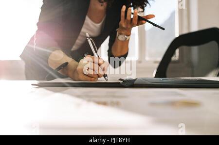 Woman entrepreneur making notes standing at her table in office while holding a cell phone in hand. Close up of a businesswoman writing in diary leani Stock Photo