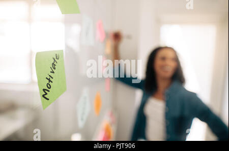 Blur image of a businesswoman pasting sticky notes on glass wall with focus on a post it note motivate written on it. Stock Photo