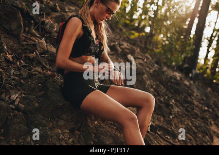 Young woman sitting by mountain trail checking her smartphone. Woman resting after trail run and checking her smart watch. Stock Photo