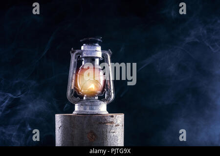 Old oil lamp burning at lumber log and foggy smoke dark night background. Rust metal lantern with corrosion. Horror and Halloween concept. Close up Stock Photo