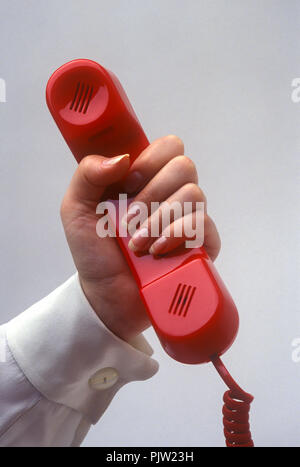 1991 HISTORICAL WOMANS HAND HOLDING UP RED LANDLINE TELEPHONE ON PLAIN RED BACKGROUND Stock Photo