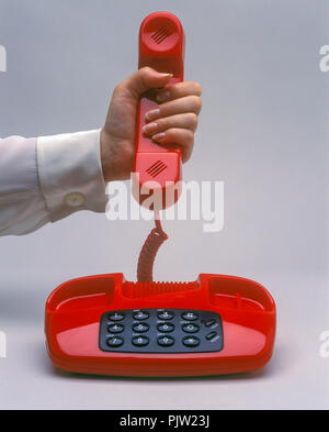 1991 HISTORICAL WOMANS HAND HOLDING UP RED LANDLINE TELEPHONE ON PLAIN RED BACKGROUND Stock Photo