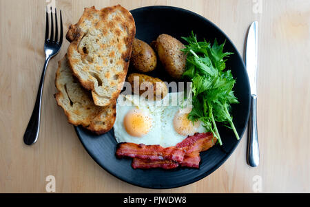 Two fried eggs, bacon, potatoes, greens and toasted rustic bread Stock Photo