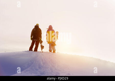 Two snowboarders male and female stands on mountain top and looks at sunset Stock Photo