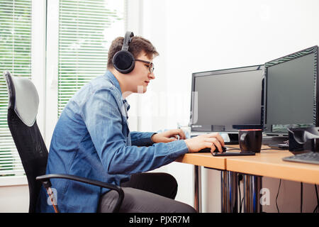 Shot of young man with headphones in casual clothes in the office, working on desktop computer. Stock Photo