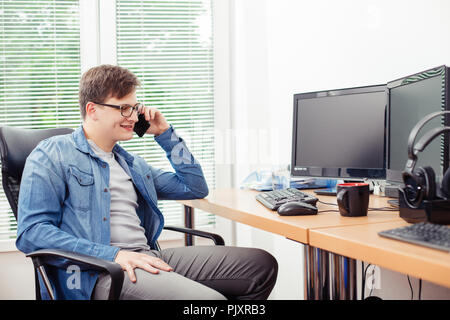 Handsome young programmer talking on the mobile phone and smiling while sitting at his working place in office Stock Photo