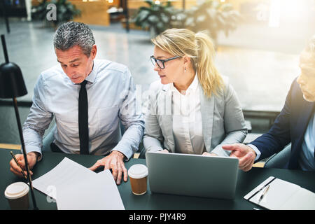 Three mature businesspeople going over paperwork and using a laptop while having a meeting together in the lobby of an office building Stock Photo