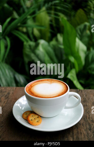 Vertical Photo of a Cup of Hot Cappuccino Coffee with Cookies Served on Wooden Table with Blurred Green Foliage in Background Stock Photo