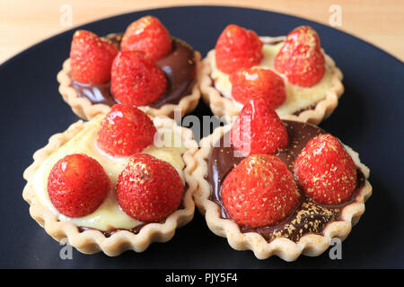 Many of mini chocolate tarts topped with fresh strawberries and edible gold powder served on black plate Stock Photo