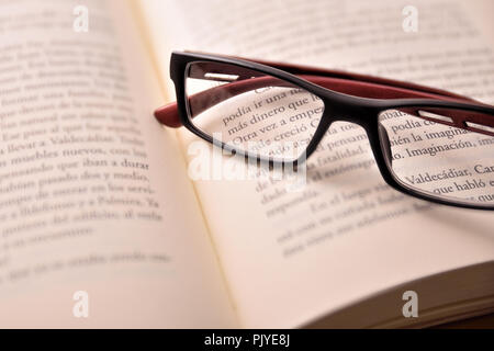 Detail of black and red reading glasses on open book.Concept need glasses to read. Elevated view. Horizontal composition Stock Photo