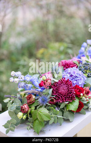 Elongated floral arrangement in vintage metal vase. table setting. Blue and red color. Gorgeous bouquet of different flowers. Park on background Stock Photo
