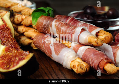 Fresh baked Italian breadsticks with Parmesan wrapped with a slice of prosciutto and olives, figs. Italian dish with antipasto on a wooden table. Stock Photo