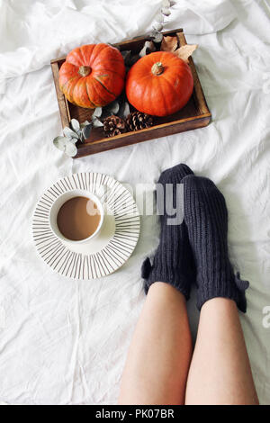 Autumn bed still life. Female legs in knitted socks and cup of coffee. Wooden tray with pumpkins, eucalyptus leaves, pine cones. White linen bed sheet background. Thanksgiving, Halloween. Flatlay, top. Stock Photo
