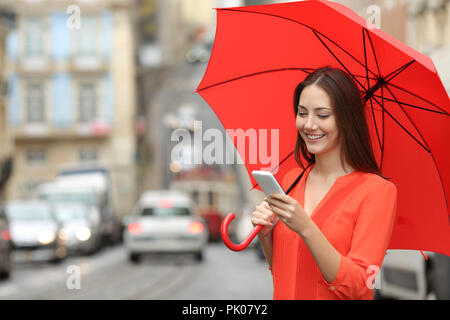 Happy woman using a smart phone holding an umbrella under the rain in the street Stock Photo