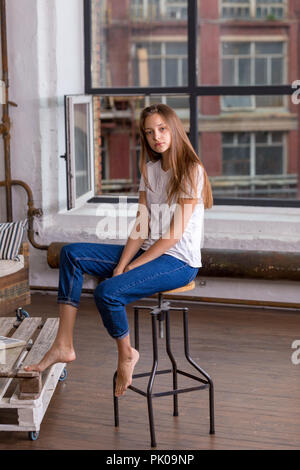 Beautiful girl with brown hair and blue eyes in white shirt and blue jeans sitting on a chair near the window  at a nice apartment Stock Photo