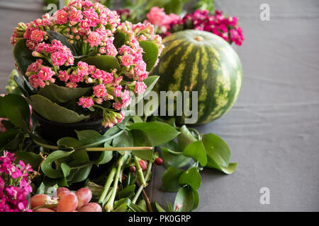 A close-up, side and birds eye view of a floral arrangement of colourful pink bunch in foreground with an enticing round watermelon & grapes. Stock Photo