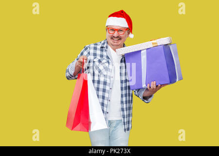 Happy middle aged man in casual style and red new year santa cap, standing and holding shopping packages and gift boxes with toothy smile, looking at  Stock Photo