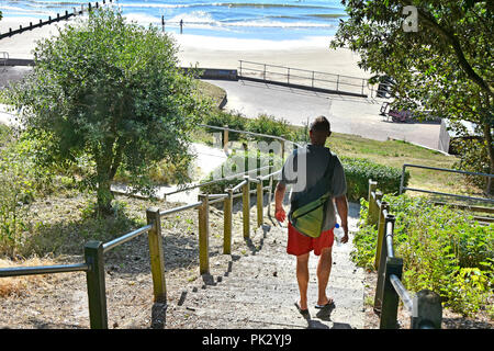 Back view of man in red shorts walking down steps towards early morning people on sandy family seaside holiday beach Frinton Essex coast England UK Stock Photo