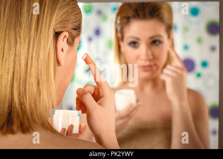 Beautiful young woman applying cream on her face in front a mirror in the bathroom at home. Stock Photo