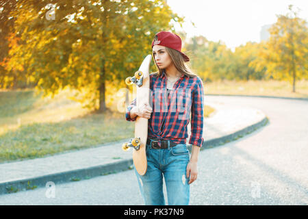 Pretty woman in shirt and jeans holding a skateboard. Beautiful portrait outdoors autumn evening at sunset Stock Photo