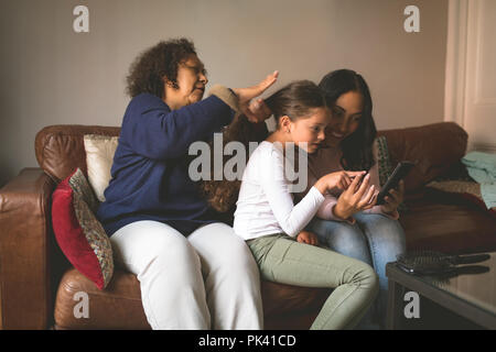 Grandmother is making hairstyle of granddaughter while using mobile with her mother Stock Photo