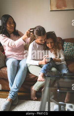 Daughter using mobile phone while mother making her hairstyle Stock Photo