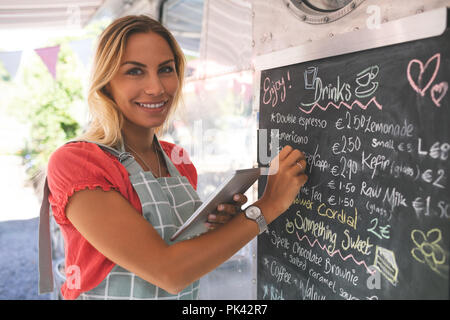 Female waitress writing menu on menu board while using digital tablet Stock Photo