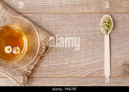 Glass cup of green tea with dry chamomile flowers in spoon on wooden background. Top view Stock Photo