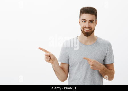 Waist-up shot of enthusiastic and charismatic handsome sportsman with beard and white pleasant smile pointing left with both fingers, smiling excited suggesting cool copy space against gray background Stock Photo