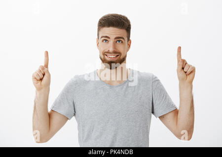 Waist-up shot of handsome masculine man with blue eyes and beard smiling joyfully and self-assured raising hands looking and pointing up delighted and happy standing pleased over gray background Stock Photo